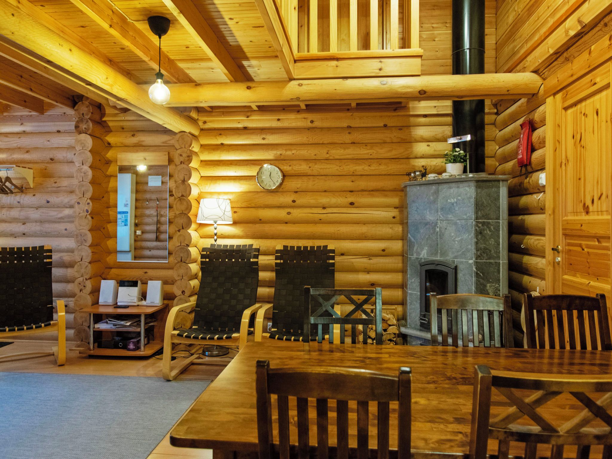 A log cabin interior with a wooden dining table, chairs, a fireplace, and a clock on the wall.