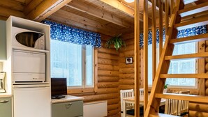 Interior de cabaña con kitchenette, paredes de madera, zona de comedor con mesa y sillas, y escalera de madera.