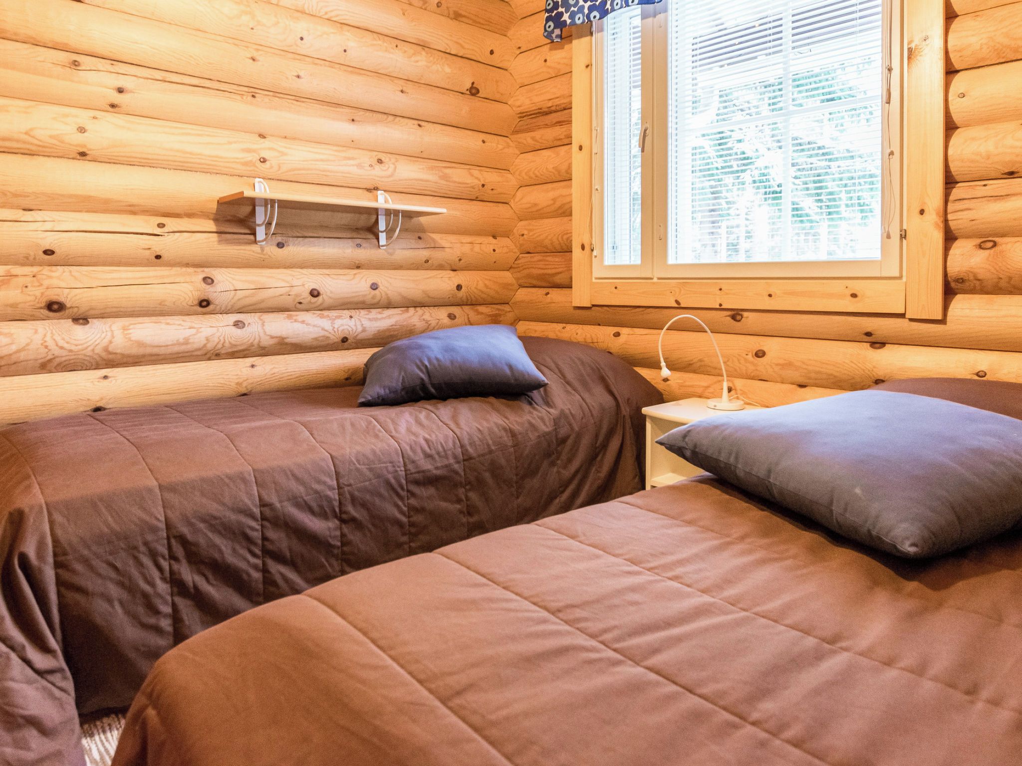 A log cabin room with two beds, a window with blinds, and a small table with a lamp.