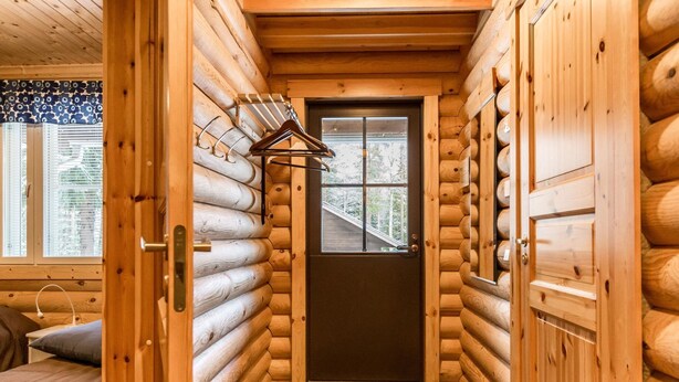 Interior de una cabaña de troncos con una puerta de madera, una cama con almohada y una ventana con persianas.