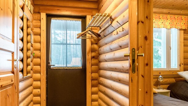 Ein Holzblockhaus-Interieur mit einer Holztür, einem kleinen Fenster mit Vorhängen, einem Bett mit Kissen und einem Nachttisch mit Lampe.