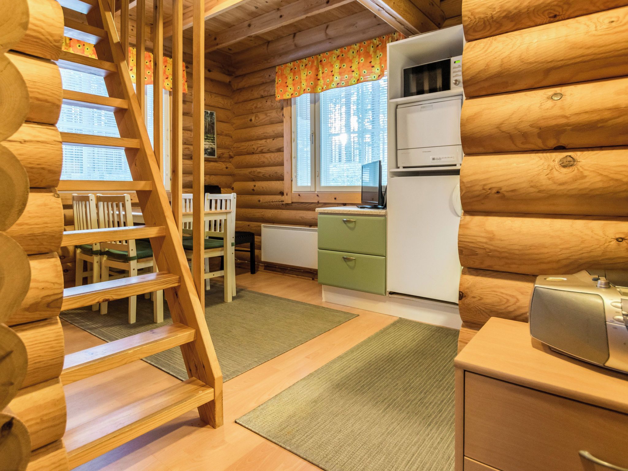 Ein Holzblockhaus-Interieur mit einer Holztreppe, einem Essbereich mit Stühlen, einer Küche mit Kühlschrank und einem Fenster mit Vorhängen.