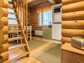 Interior de una cabaña de troncos con escalera de madera, comedor con sillas, cocina con refrigerador y ventana con cortinas.