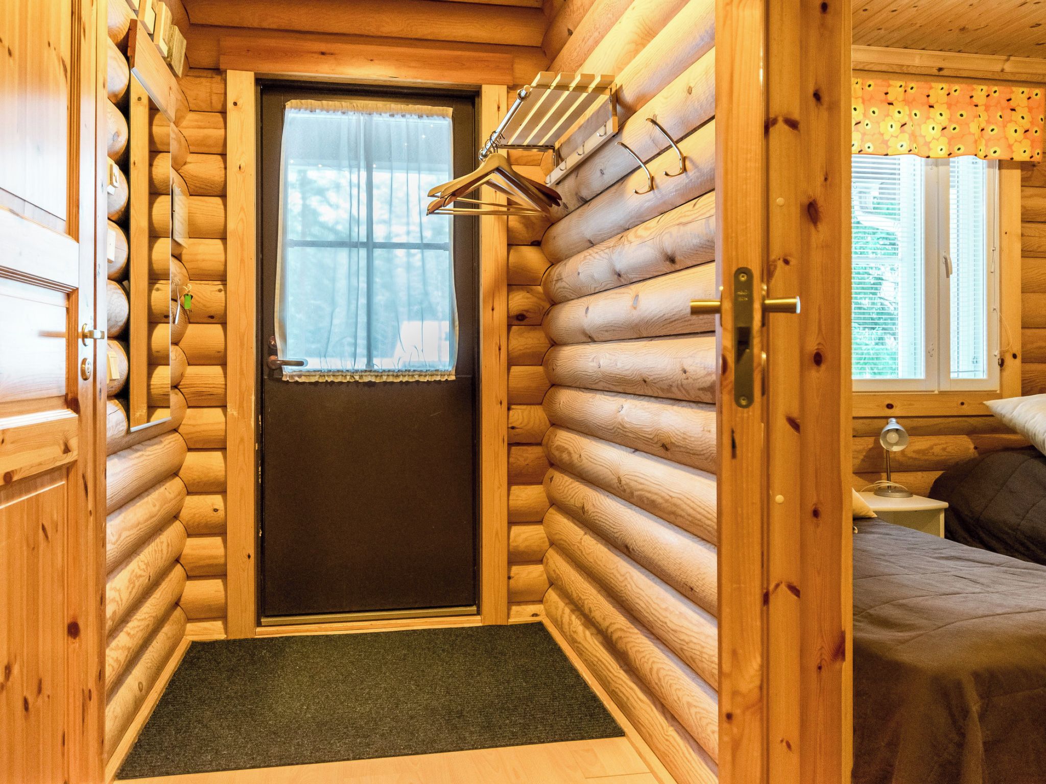 L’intérieur d’une cabane en rondins, avec une porte donnant sur une chambre, une petite fenêtre avec un rideau et un porte-manteau avec des cintres.