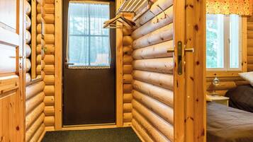 L’intérieur d’une cabane en rondins, avec une porte donnant sur une chambre, une petite fenêtre avec un rideau et un porte-manteau avec des cintres.