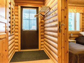 Interior de una cabaña de troncos con una puerta que da a un dormitorio, un ventanal pequeño con cortina y un perchero con ganchos.