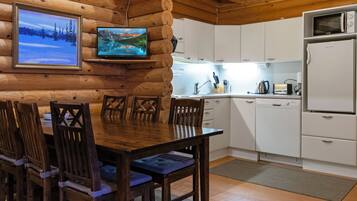 A wooden cabin interior with a dining table, chairs, a television mounted on the wall, a kitchen area with cabinets and appliances, and a framed picture on the wall.