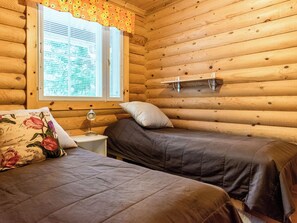 Habitació d'una cabana de fusta amb dos llits, una finestra amb persianes, una tauleta de nit i una làmpada.