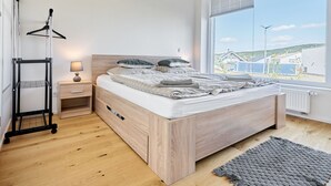 A bedroom with a wooden bed, a nightstand, a lamp, a hanging clothes rack, and a large window with a view of buildings and greenery. - Ore house by Interhome (Loucna pod Klínovcem)