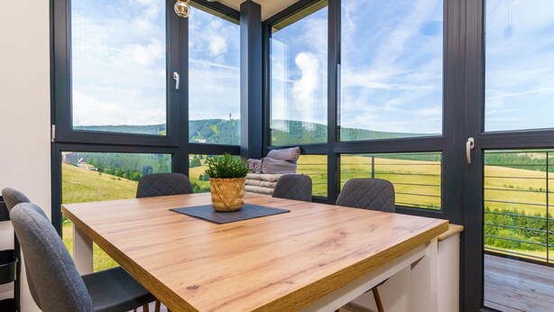 Un coin repas avec une table en bois, quatre chaises et une vue sur des champs verdoyants, visible à travers de grandes fenêtres.
