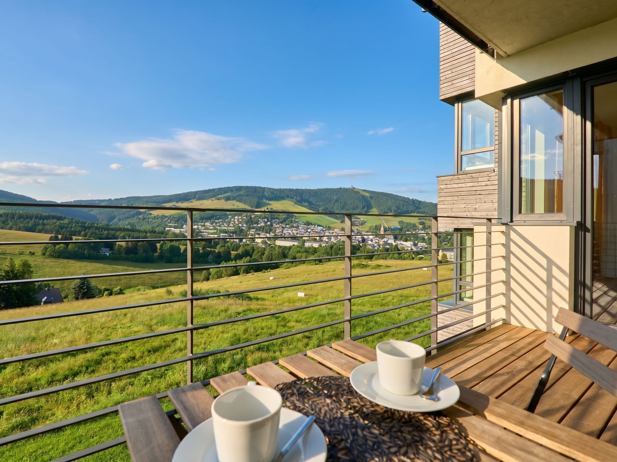 Un balcon donnant sur la ville et les collines, une table avec une tasse et une soucoupe, et un banc en bois.