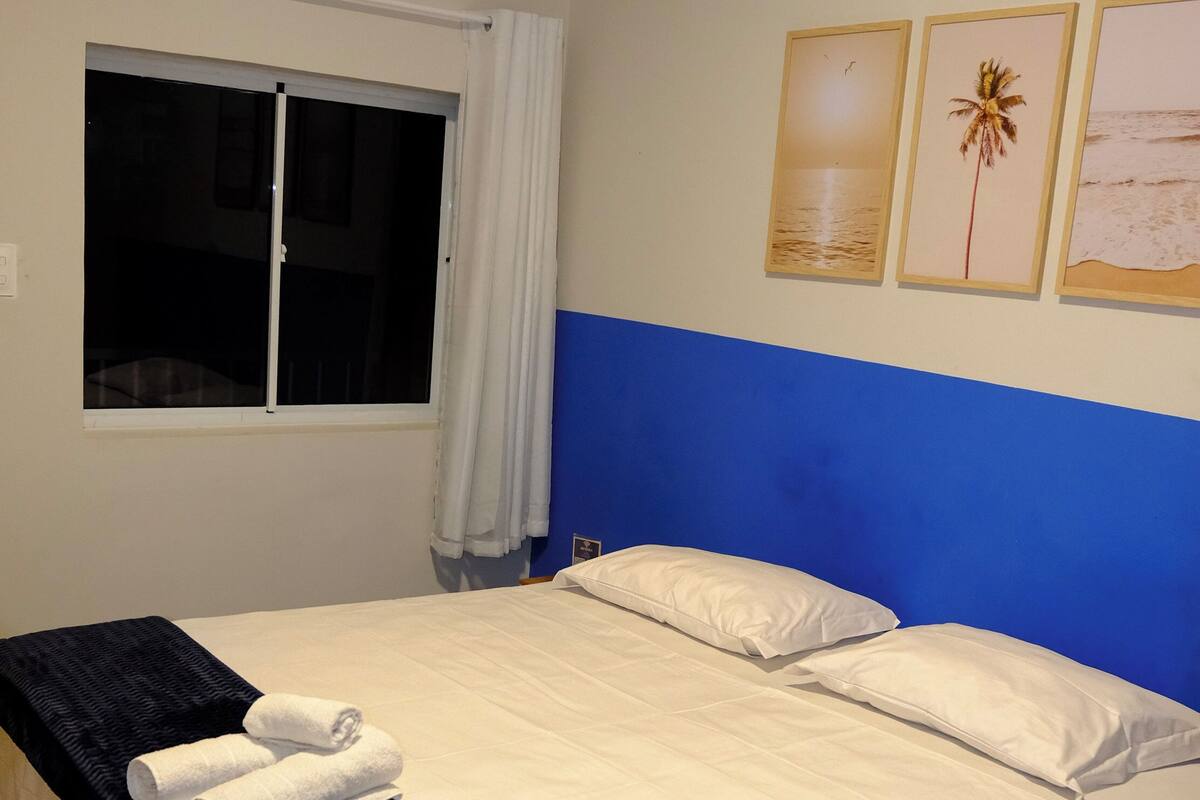Una cama bien tendida con sábanas blancas y un edredón negro, una ventana con cortinas blancas y un aire acondicionado de pared.