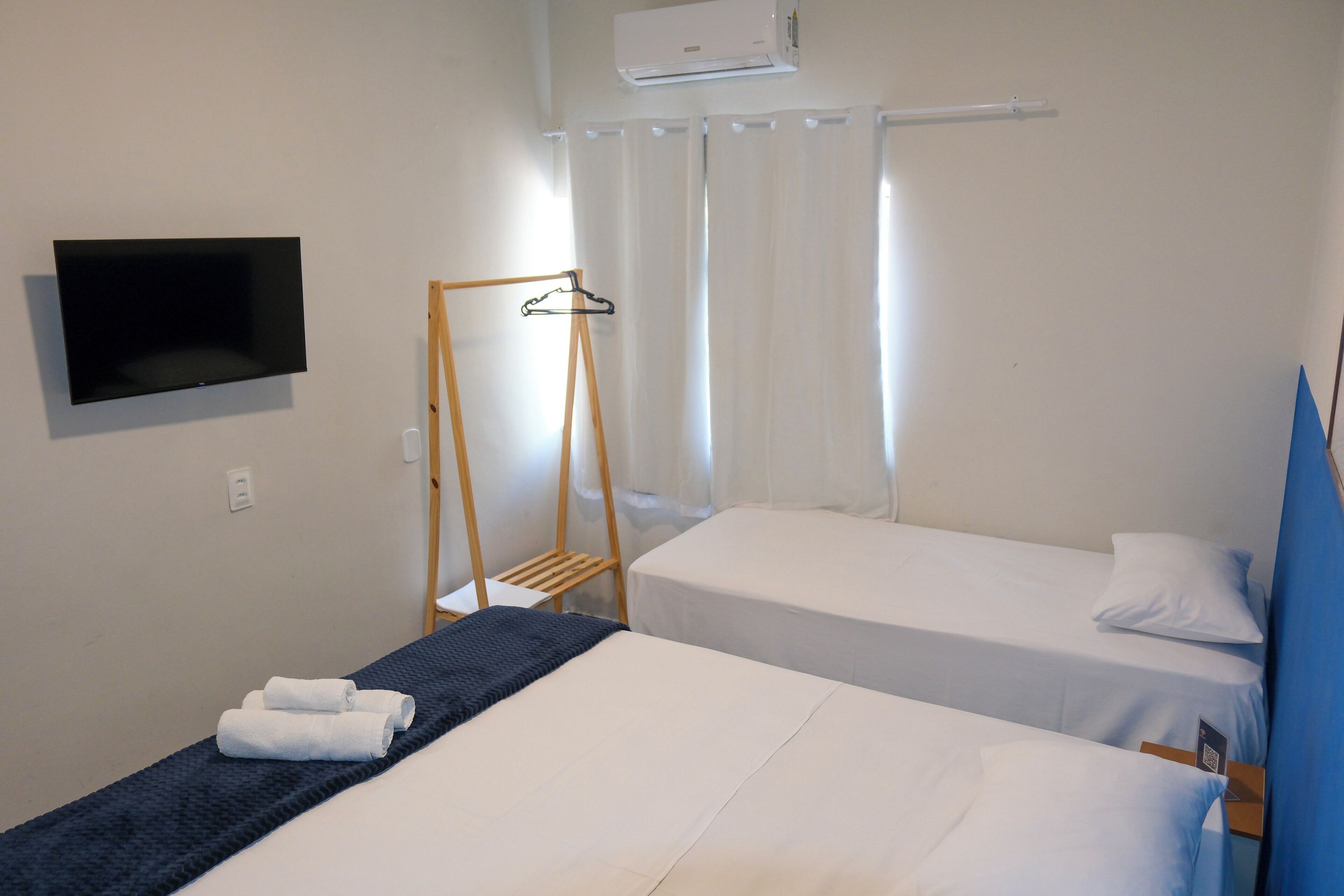 Quarto de hotel com duas camas de solteiro, televisão fixada na parede, um cabide de madeira e uma janela com cortinas.