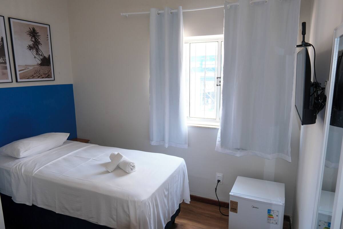 Una habitación de hotel con una cama, un armario blanco pequeño y una ventana con cortinas.