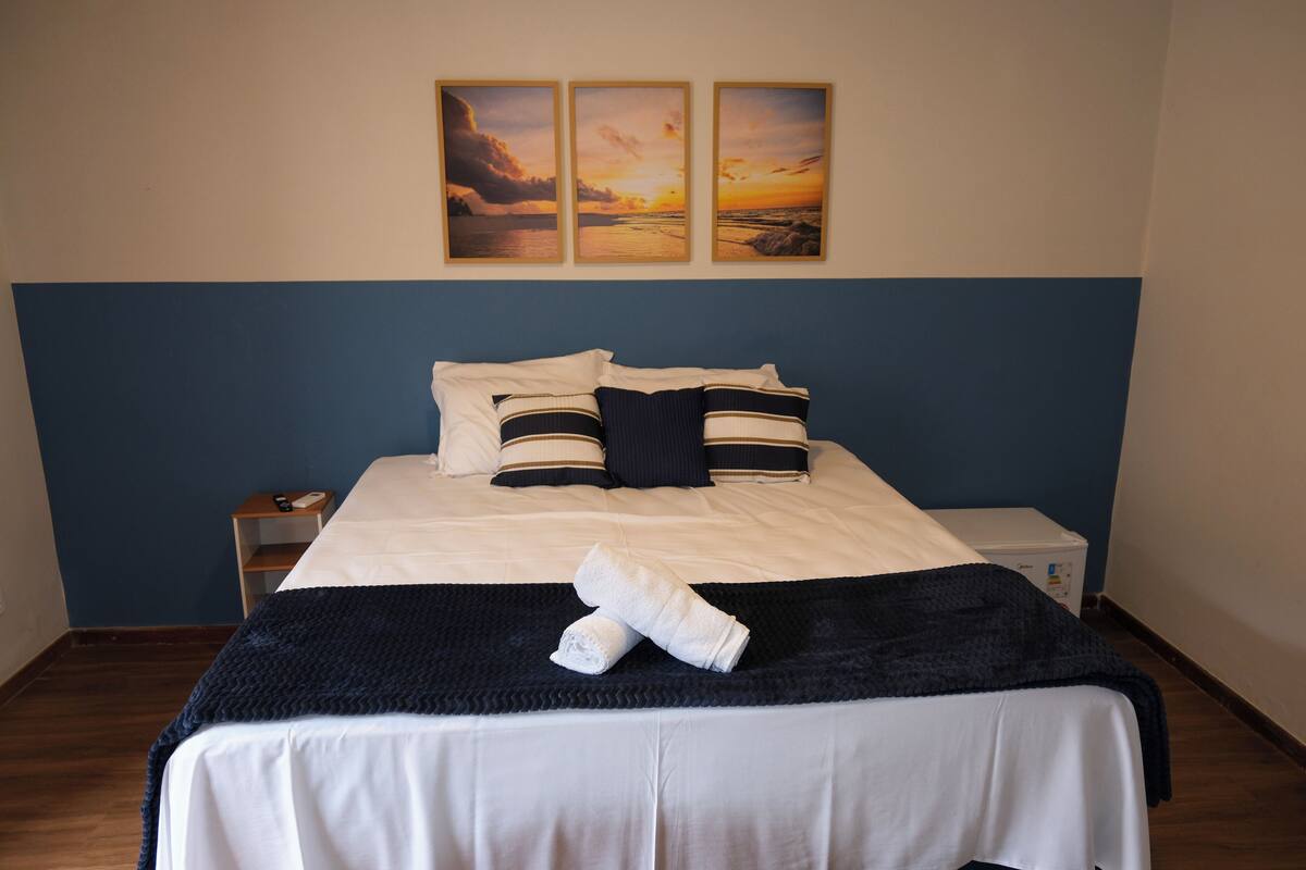 Una cama bien tendida con ropa de cama blanca y un cabecero azul oscuro. Hay tres cuadros enmarcados en la pared sobre la cama.
