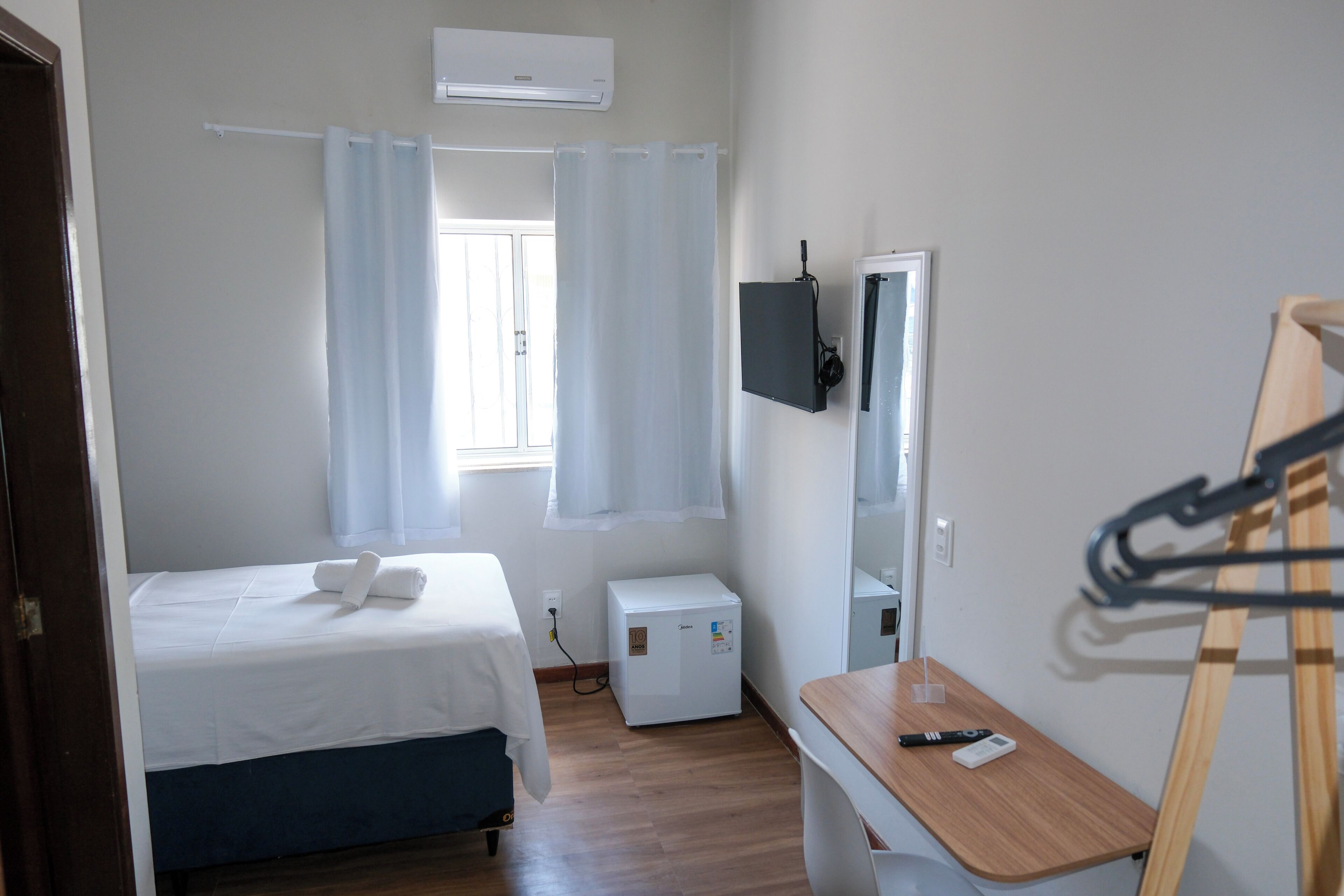 A small, clean room with a bed, a desk, a mirror, and a window with curtains.