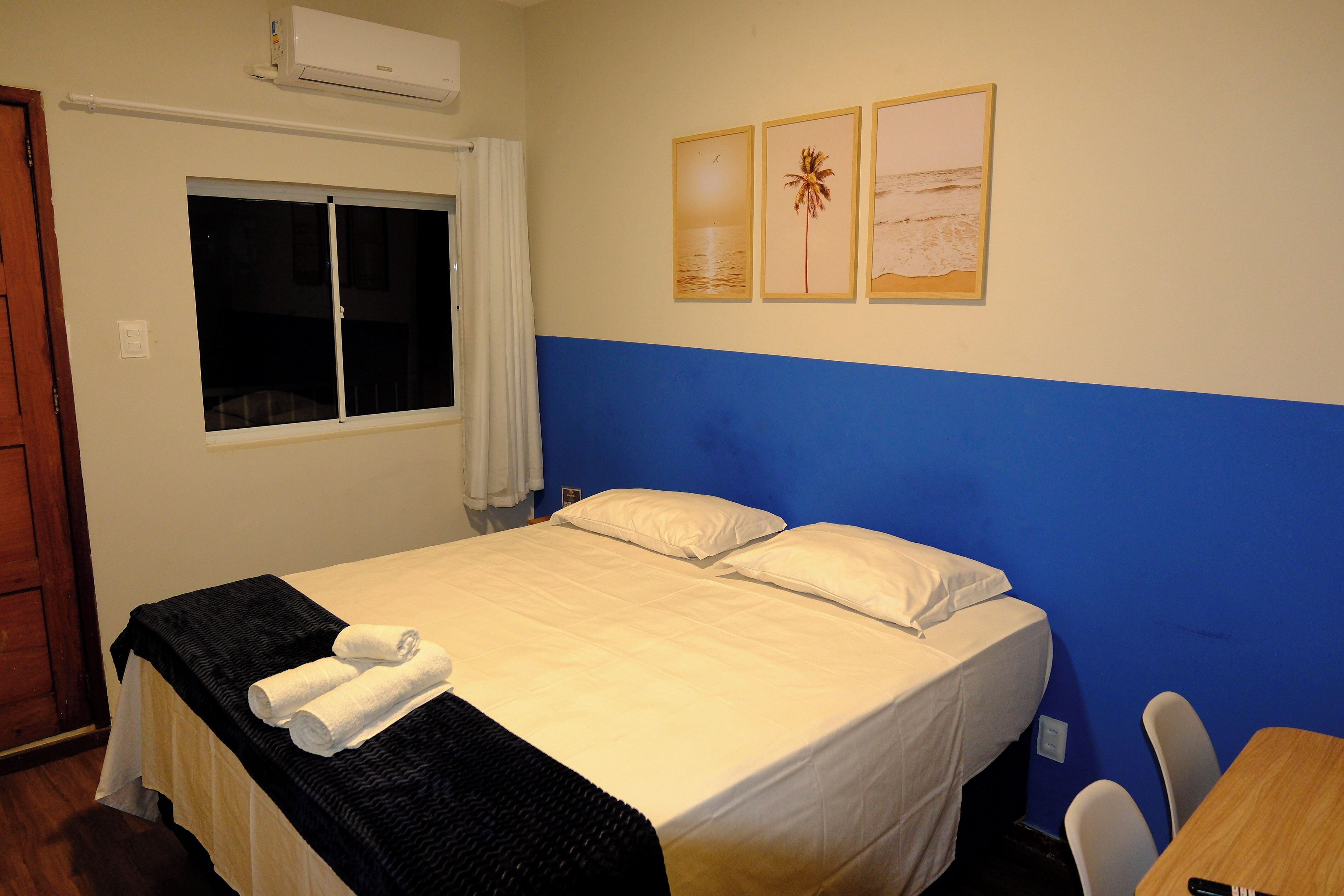 Una cama individual con sábanas blancas, una manta negra y dos toallas enrolladas. Hay una ventana con cortinas blancas, un aire acondicionado de pared y tres cuadros con imágenes de un atardecer, una palmera y una playa.