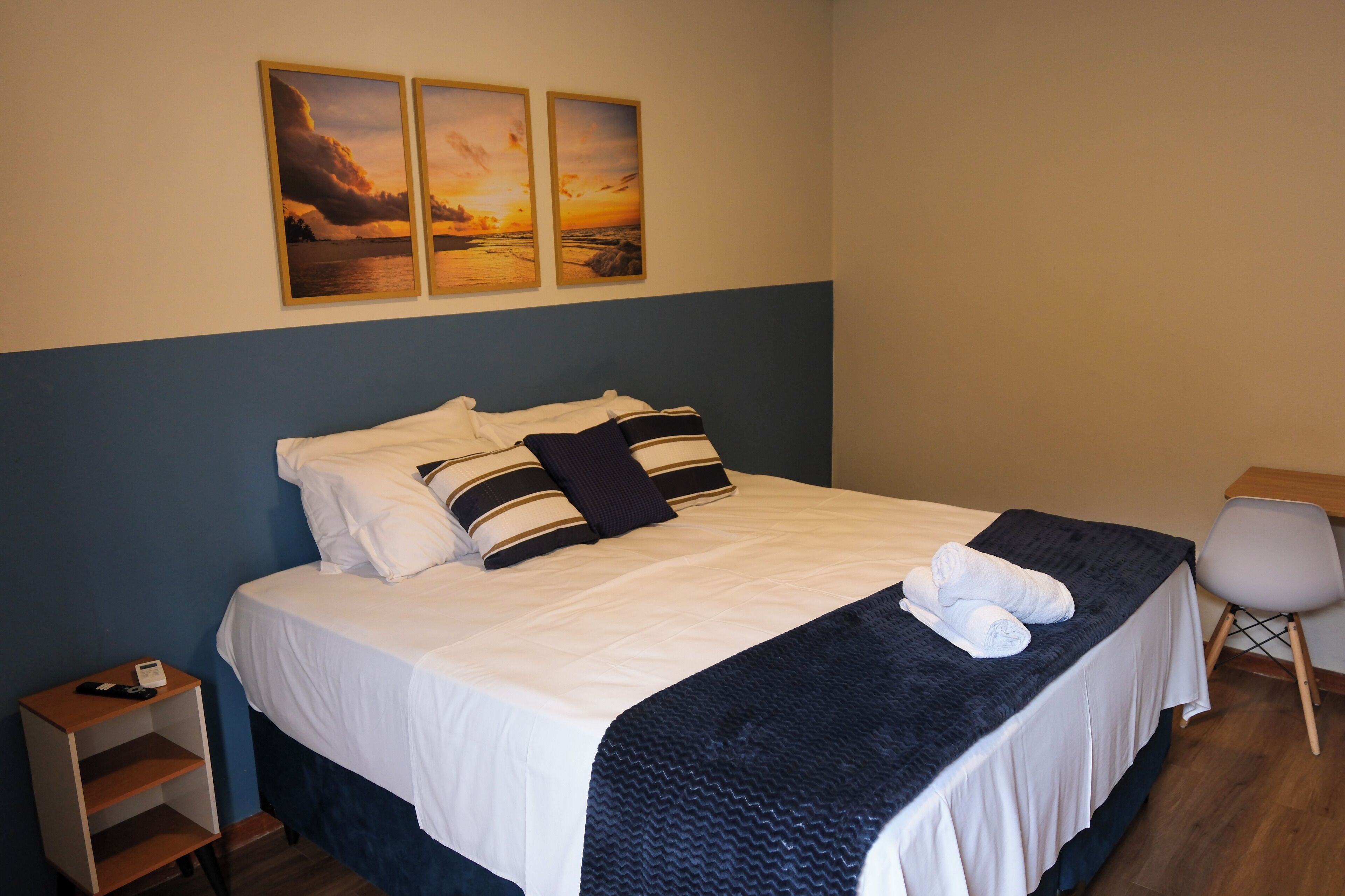 A neatly made bed with white and striped pillows, a dark blue blanket, and a pair of towels on the bed. There are two paintings on the wall above the bed depicting a sunset over water.