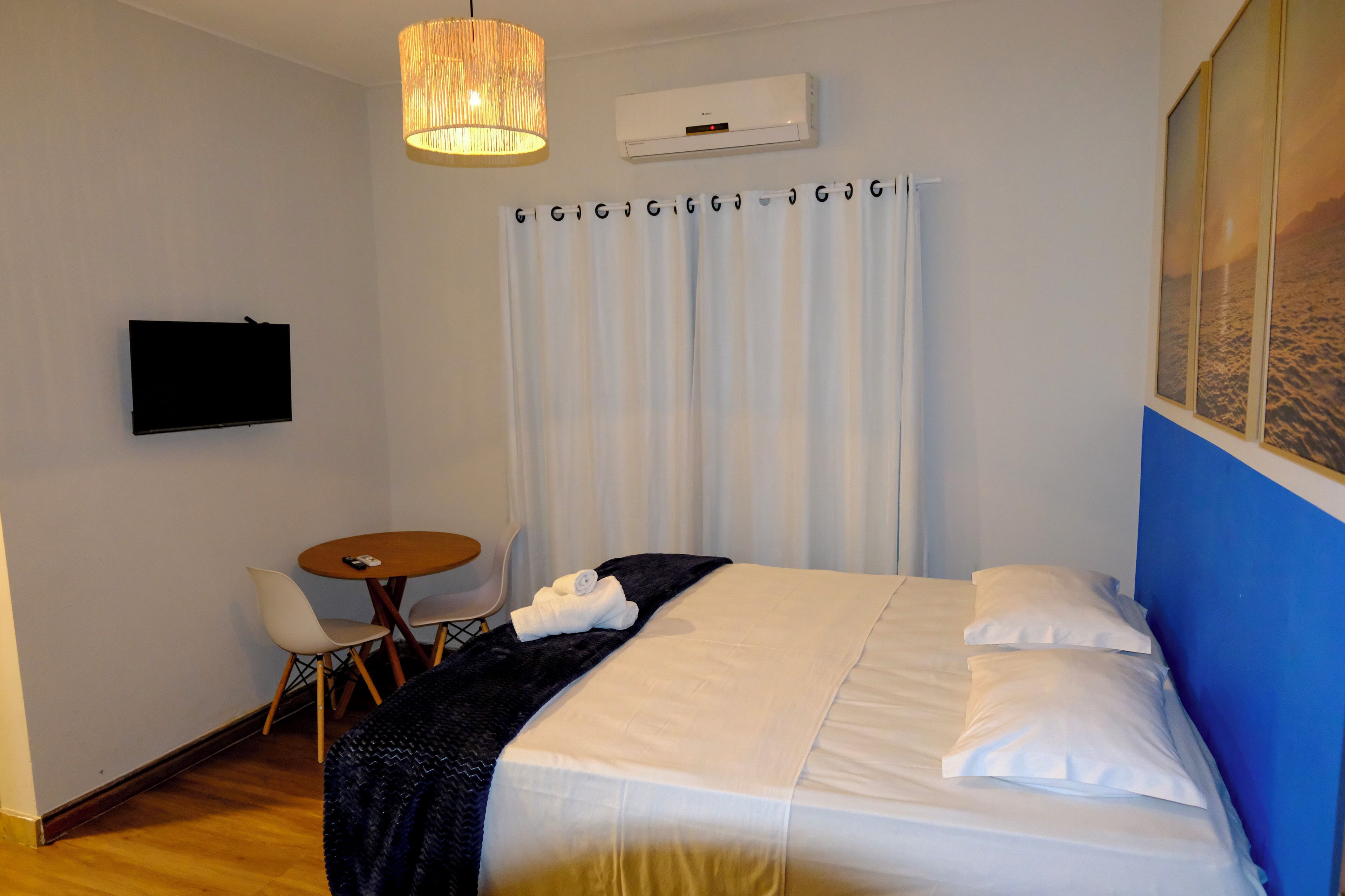 A hotel room with a bed, a TV mounted on the wall, a small round table with chairs, and a window with curtains.