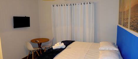 A hotel room with a bed, a TV mounted on the wall, a small round table with chairs, and a window with curtains.