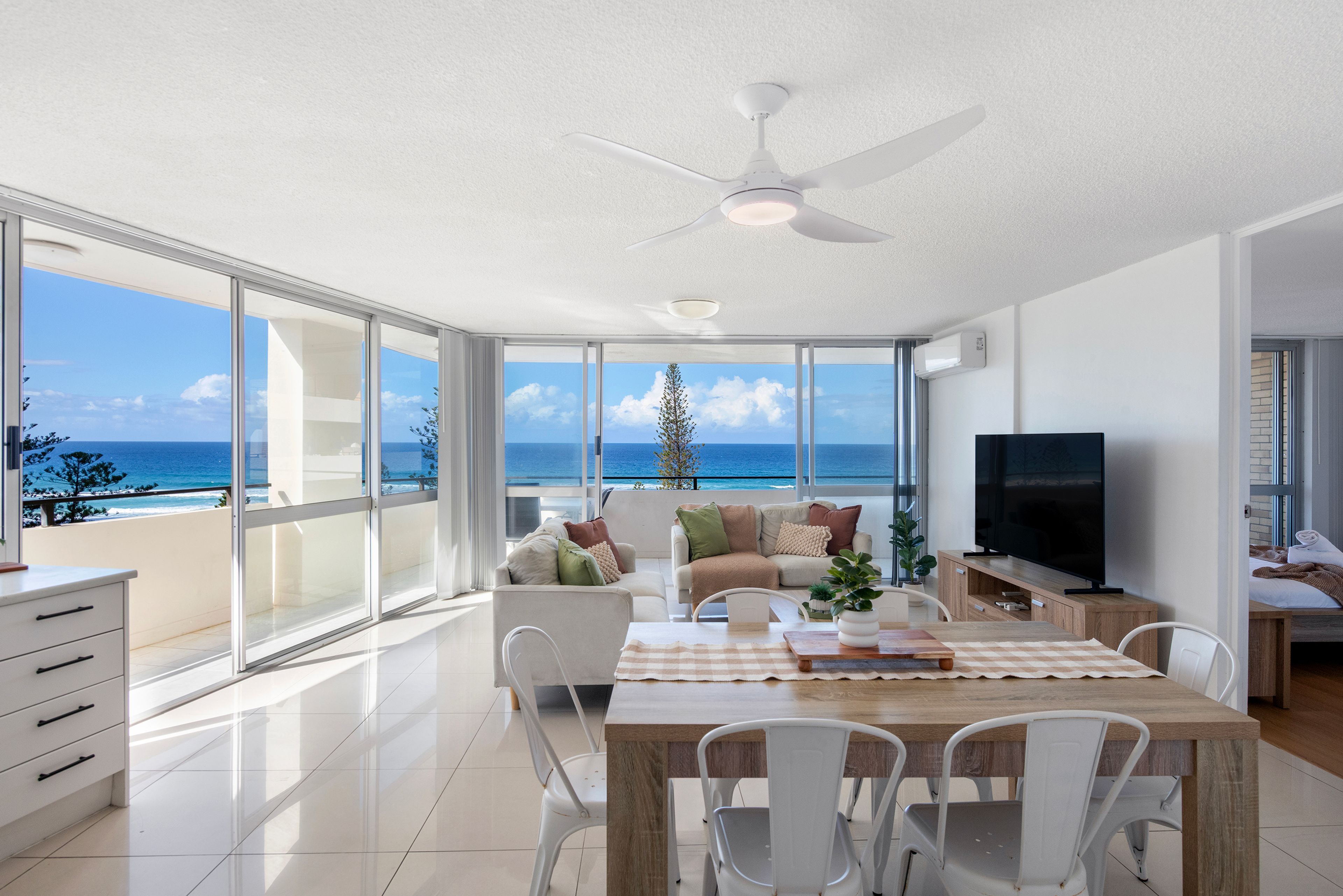 A spacious living room with a large TV, a dining area, and a balcony with a view of the ocean.