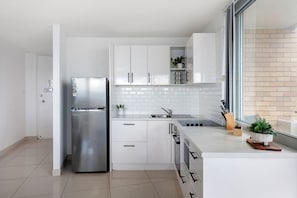 Et moderne køkken med hvide skabe, et rustfrit stål køleskab, indbygget ovn og et vindue med naturligt lys.