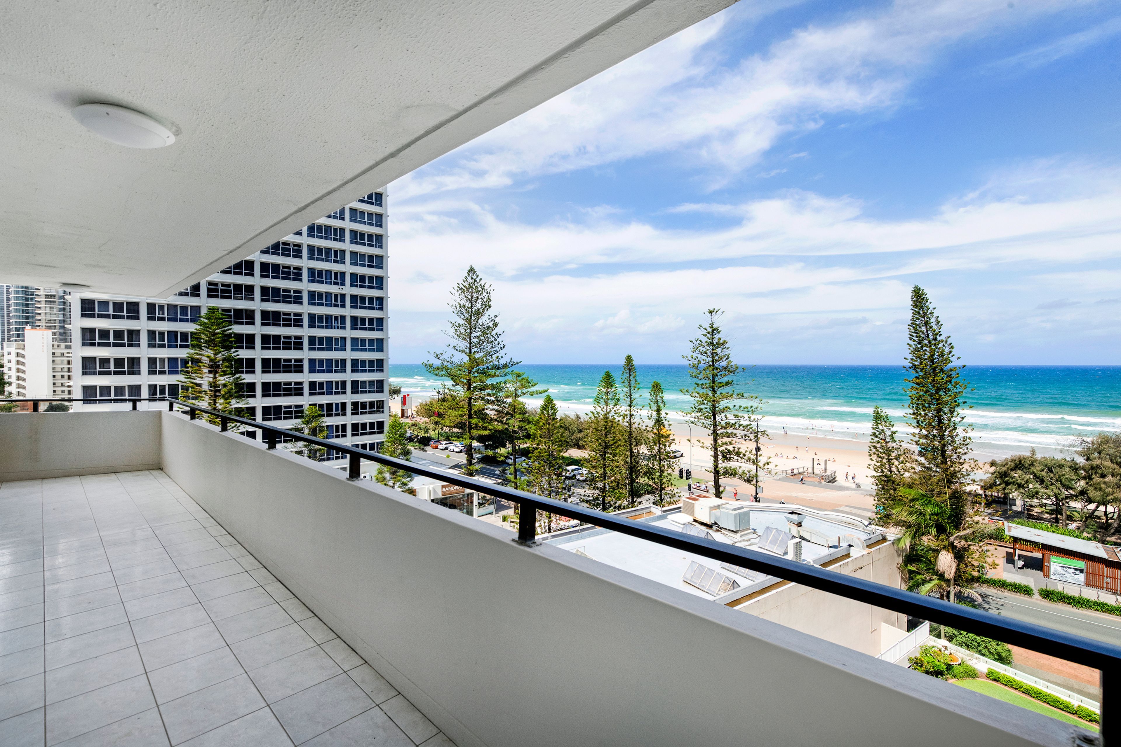 A balcony with a view of a beach and ocean, a parking area, and a modern building in the background.