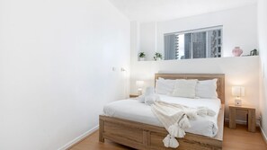 Une chambre avec un lit en bois, du linge de lit blanc, deux lampes de chevet, une fenêtre donnant sur des immeubles et une étagère avec des plantes.