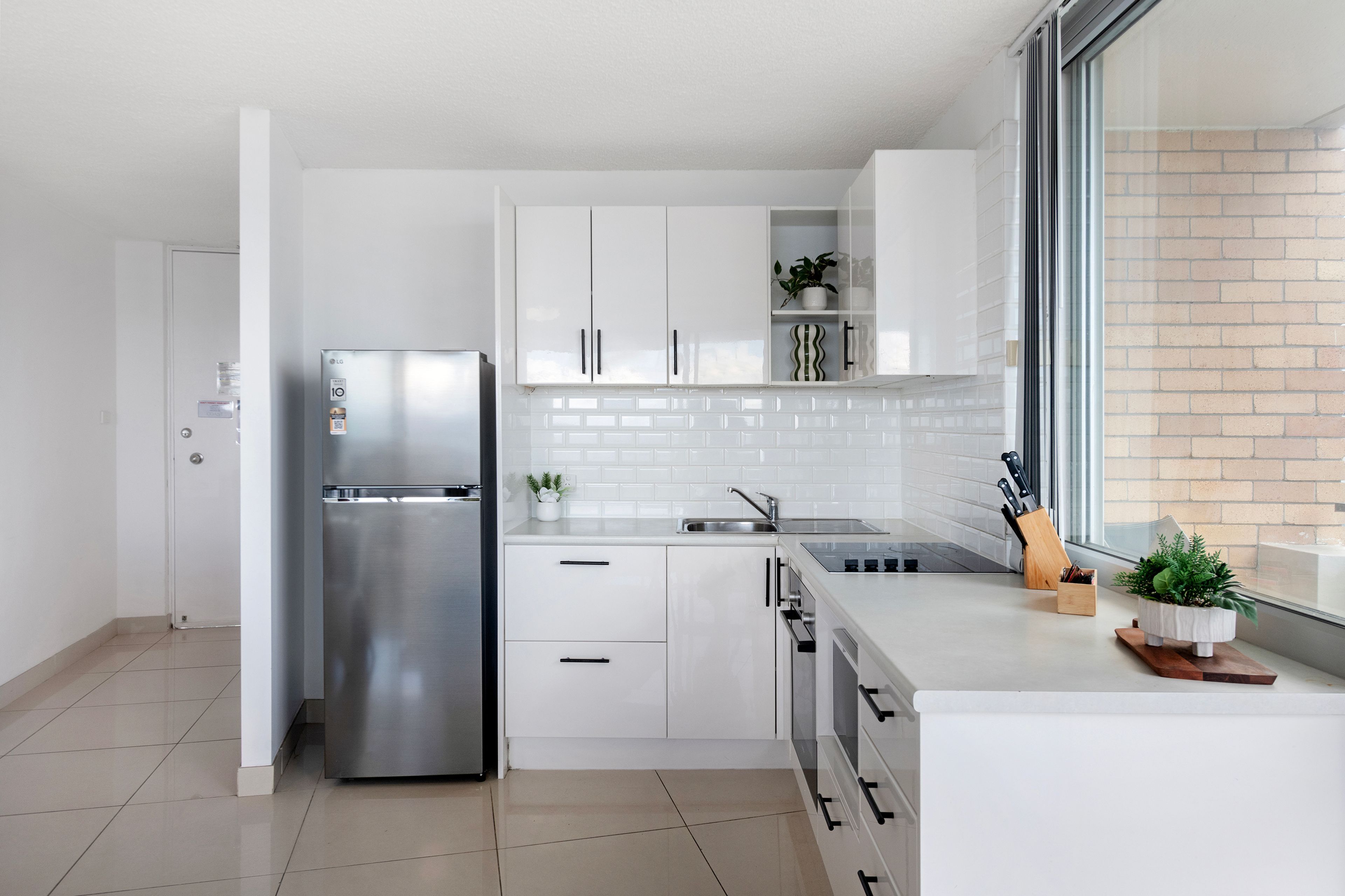 A modern kitchen with white cabinetry, a stainless steel refrigerator, a built-in oven, and a window with a view of the outside.