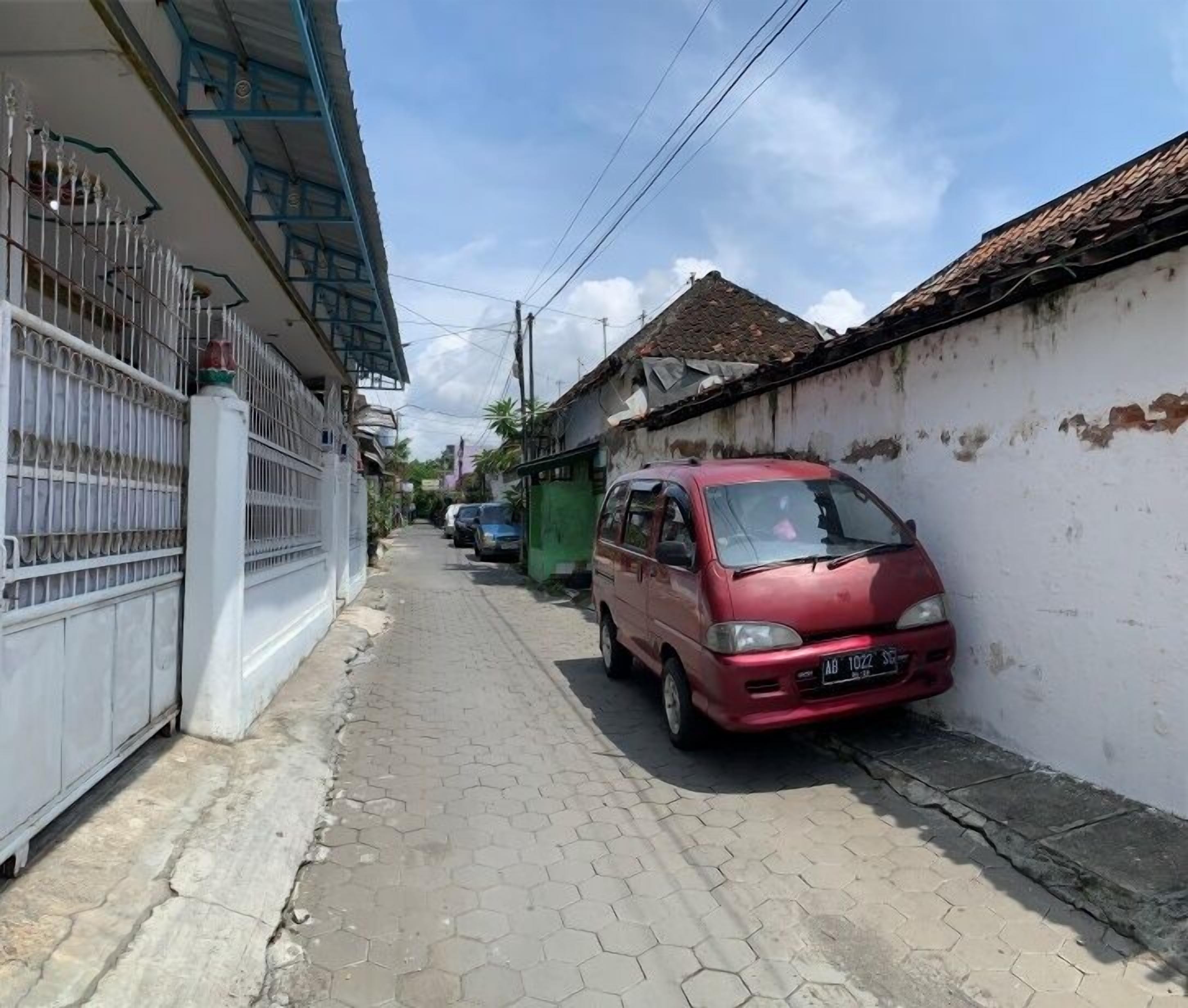 A narrow street with a red car parked on the right, a white fence on the left, and a building with a blue roof in the background.