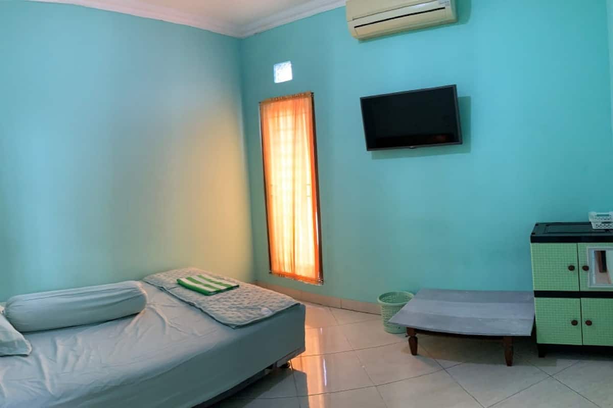 A small, single-bed room with a television mounted on the wall, a window with curtains, and a green cabinet with drawers.