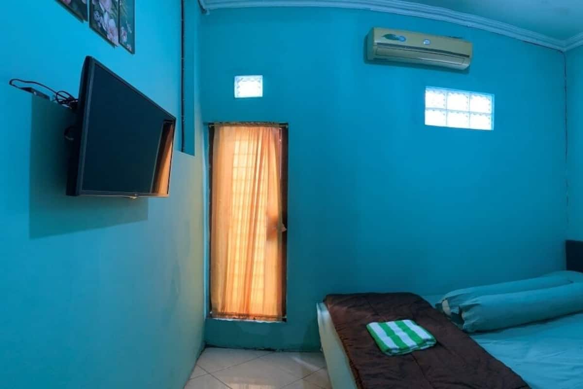 A small room with teal walls, a single bed with blue bedding, a wooden table with a green and white striped cloth, a mounted TV, and a window with curtains.