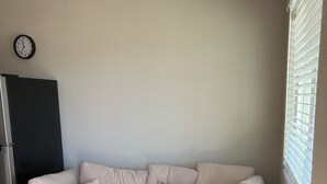 Un salon avec un canapé d’angle beige, un coussin décoratif à motifs, une horloge au mur et une fenêtre avec des stores.