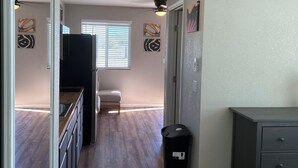 Un couloir étroit, avec une kitchenette d’un côté, une fenêtre avec des stores et une porte donnant sur une autre pièce.