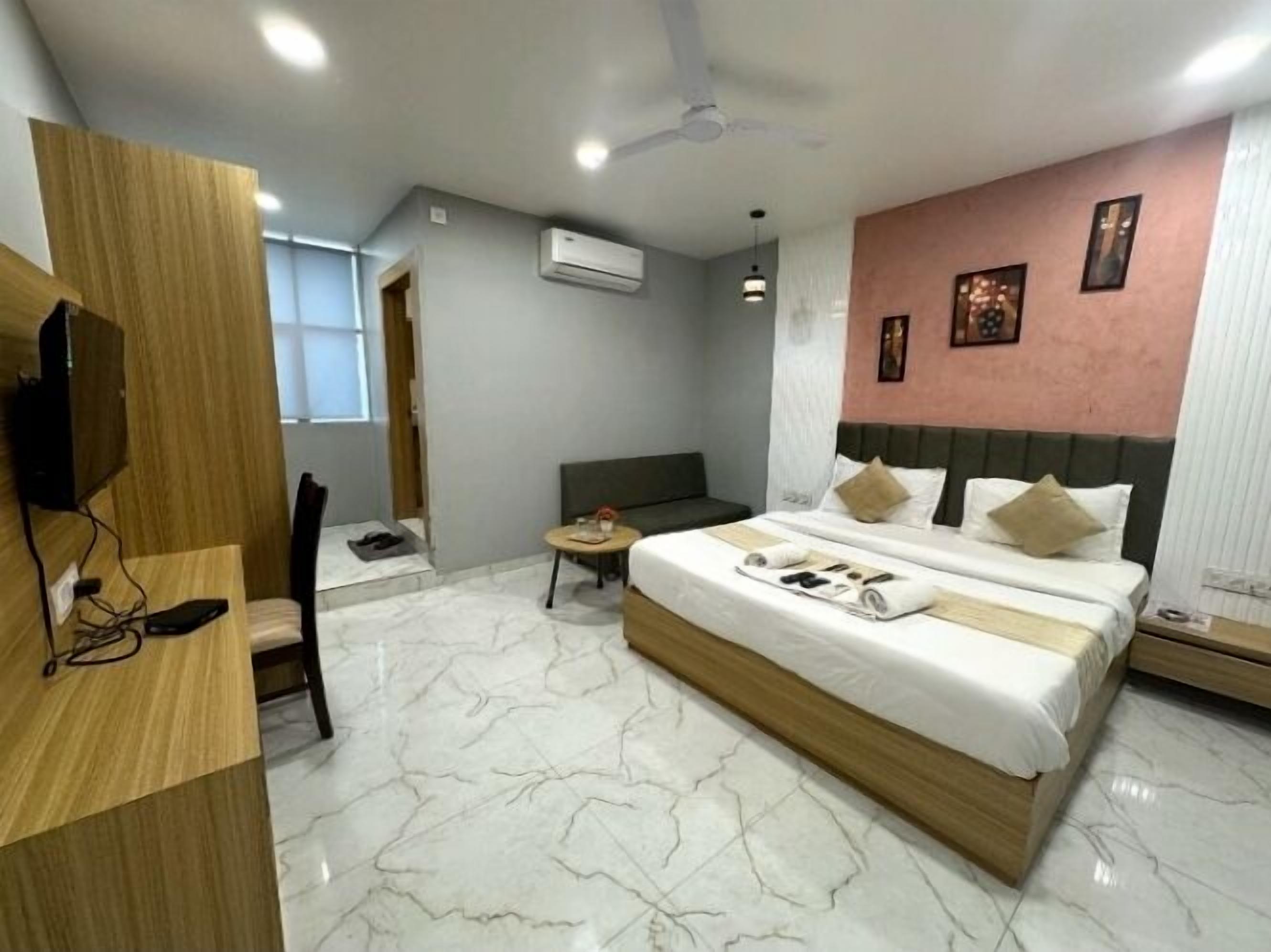 Ein modernes Hotelzimmer mit Marmorboden, einem großen Bett mit Holz Kopfteil, einem Schreibtisch mit Stuhl, einem kleinen runden Tisch und einem Deckenventilator.