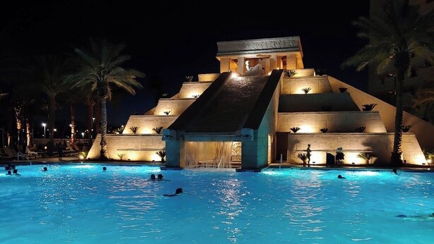 Una piscina con una estructura en forma de pirámide al fondo, palmeras y gente nadando.