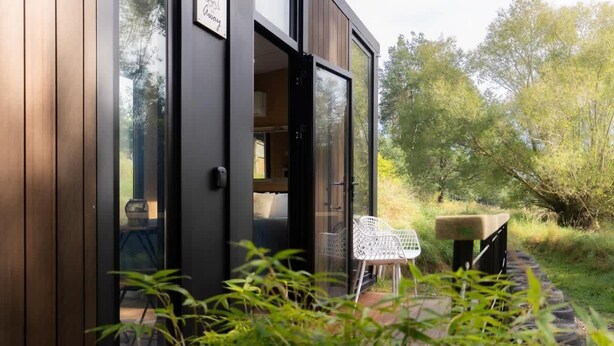 Ein modernes Gebäude mit geöffneter Glastür, dahinter ein Zimmer mit Bett und Stuhl.