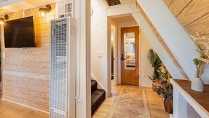 A modern interior with a wooden floor, a large TV mounted on the wall, and a staircase leading to an upper level. - Cozy A-Frame High in The Tree Tops (Crestline)