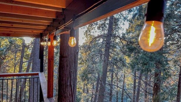 A wooden deck with hanging light bulbs and a view of a forest. - Cozy A-Frame High in The Tree Tops (Crestline)