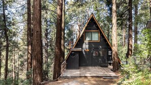 A cabin with a triangular roof, a wooden deck, and a door with a window above it, surrounded by tall trees. - Cozy A-Frame High in The Tree Tops (Crestline)