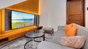 A modern living room with a large flat-screen TV mounted on a wooden wall, a glass coffee table, a grey sofa with orange and white cushions, and a wooden door. - Villa Prinz near Nusa Dua Beach (Nusa Dua)