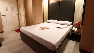 A hotel room with a bed, a bedside table with a phone and a menu stand, a door with a red mat, and a wall-mounted light.