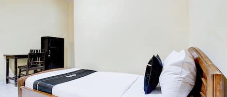 A single bed with a wooden frame, white bedding, and two pillows. A small wooden table and a black cabinet are visible in the background.
