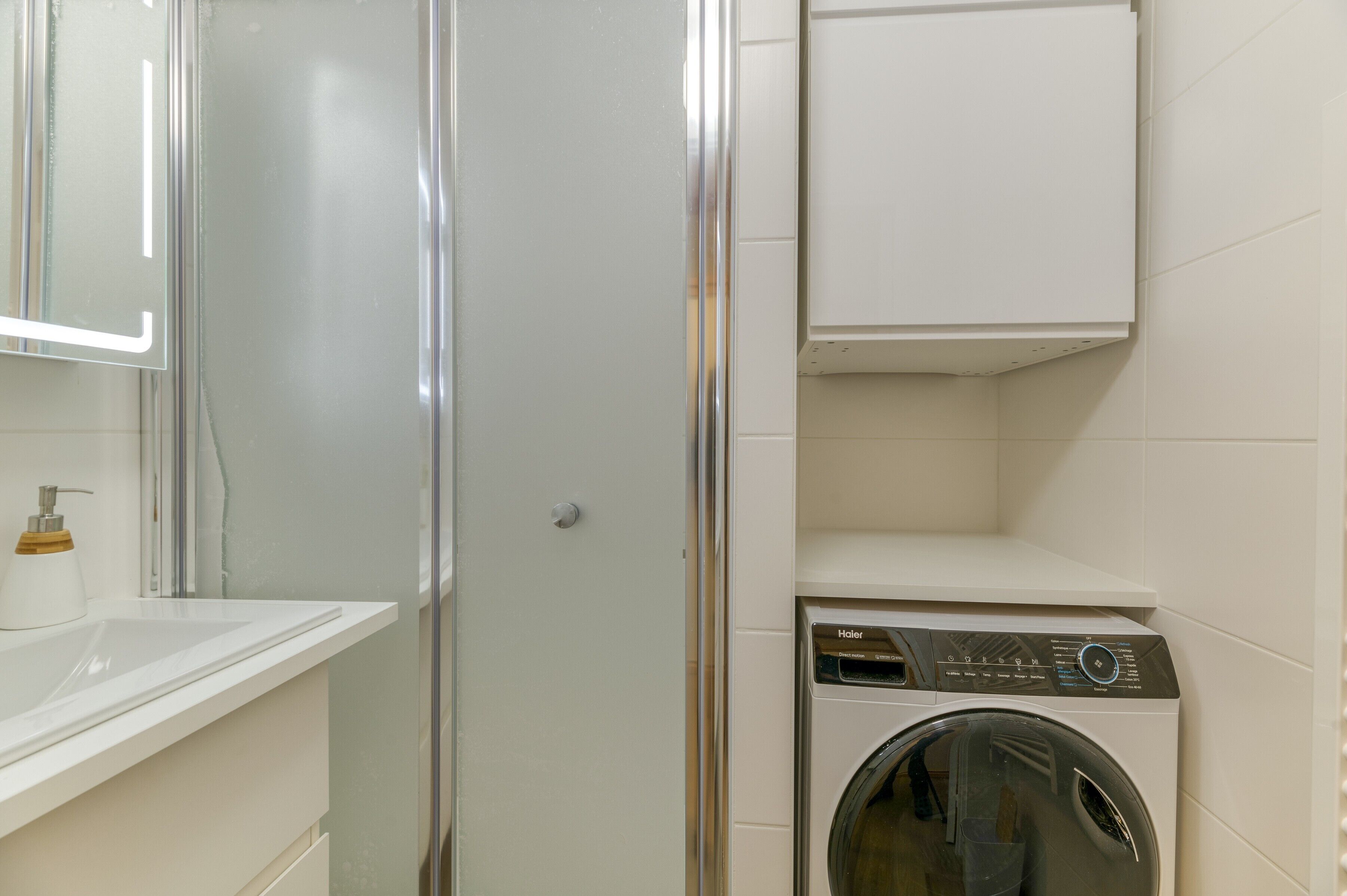 Une salle de bain avec une douche à parois de verre, un lavabo doté d’un distributeur de savon et un lave-linge à chargement frontal.