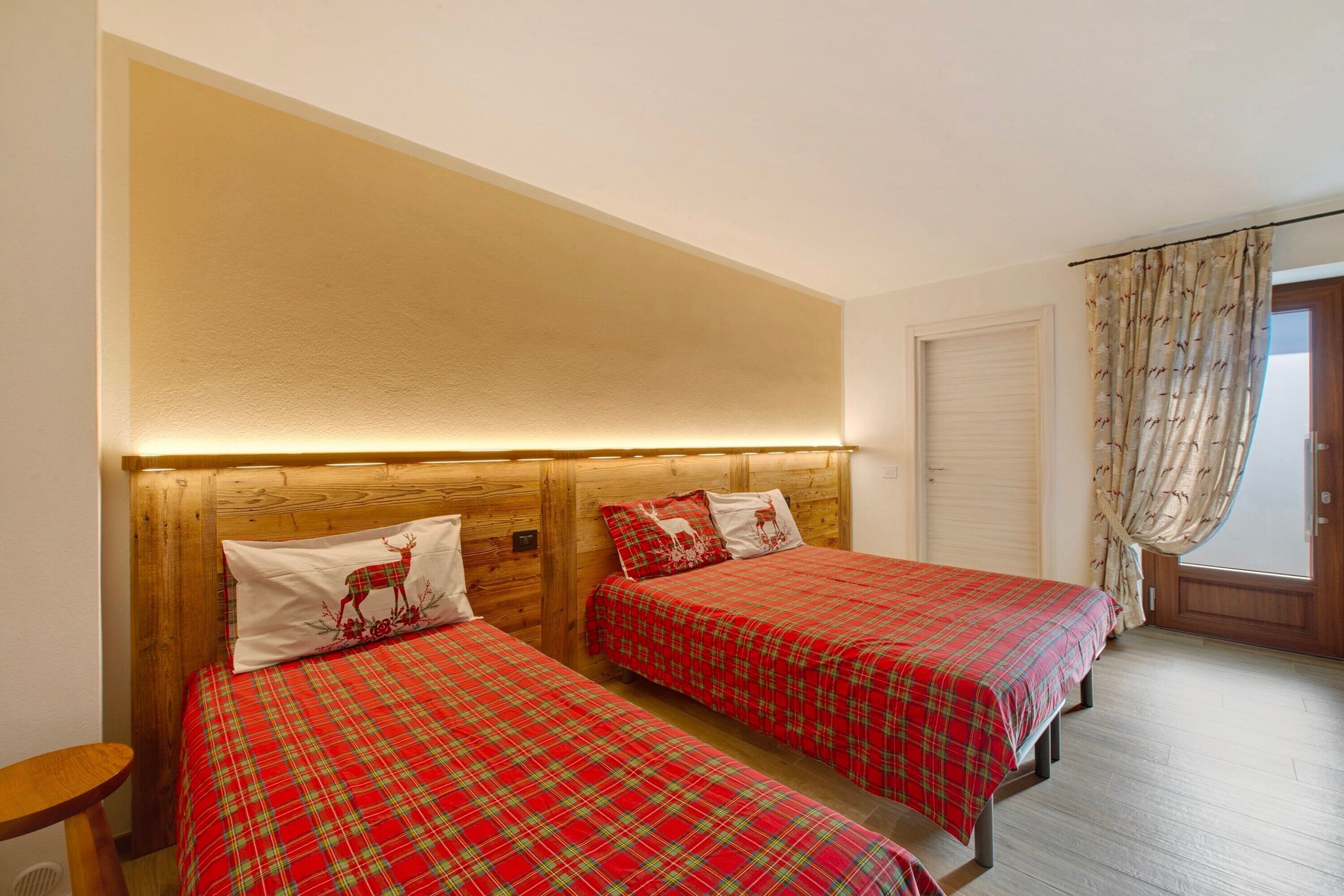 Een kamer met twee bedden, houten hoofdborden, een ronde houten kruk en een raam met gordijnen.