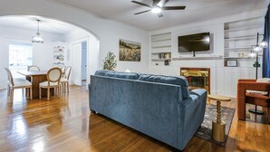 A living room with a blue sofa, wooden dining table, chairs, a fireplace, and a TV mounted above it. - Timeless Getaway in San Angelo! Fully renovated 3BR/2BA 1920s Home (San Angelo)