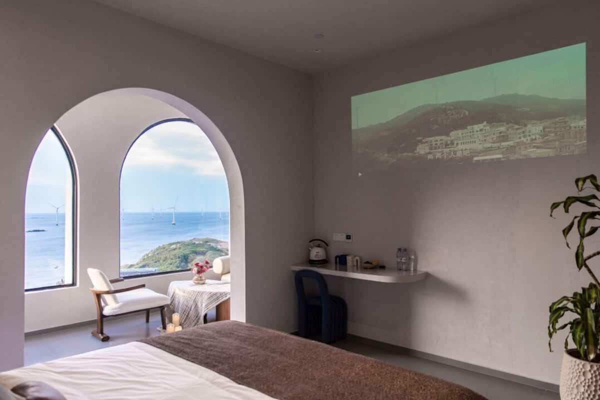 A hotel room with a large bed, a desk, and a view of the sea through arched windows.
