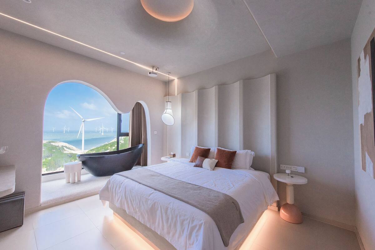 A modern bedroom with a large bed, a view of wind turbines, and a unique arched window.