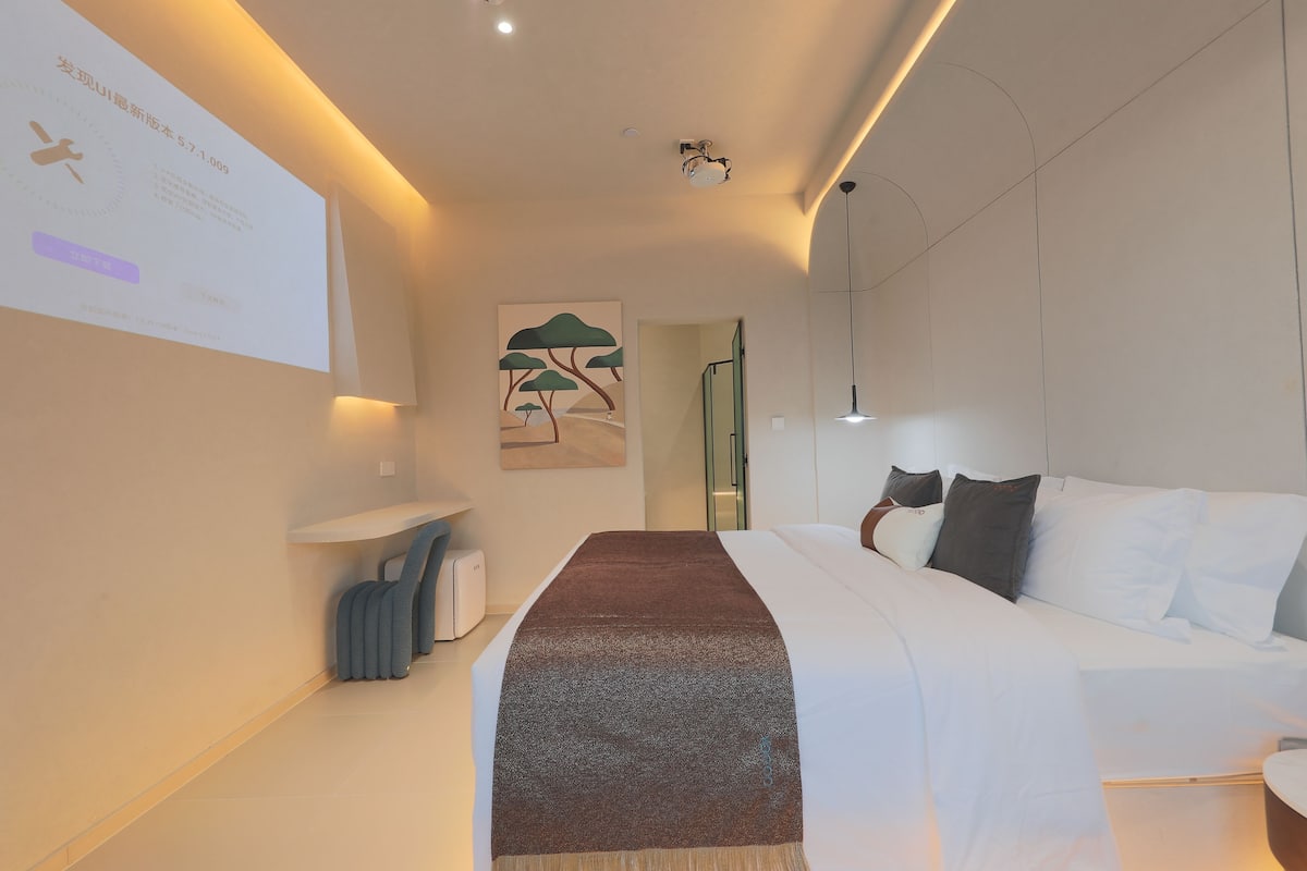 A modern hotel room with a bed, a bedside table, a wall-mounted projector, and a painting of trees.