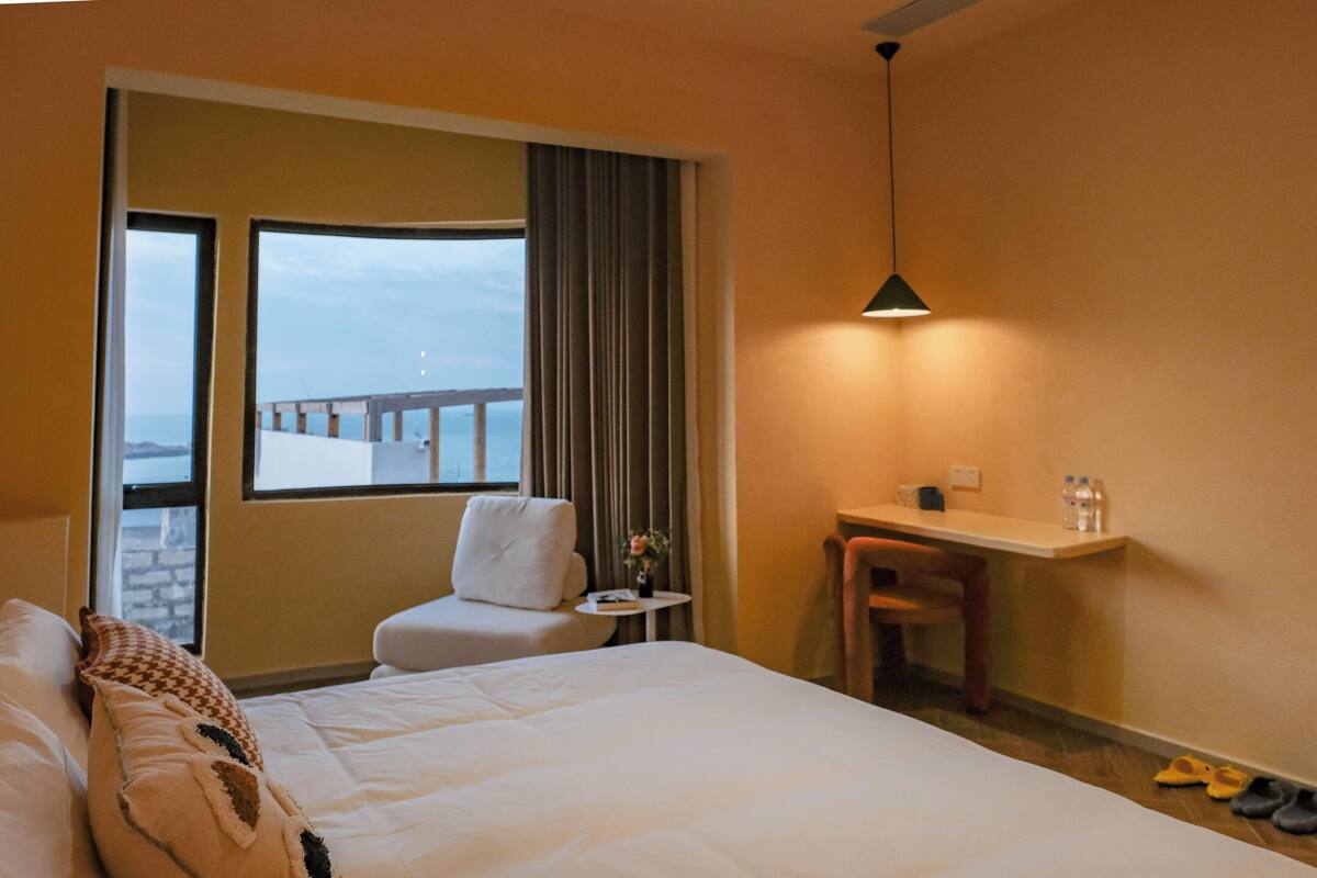 A hotel room with a bed, a desk, a chair, a small table, and a view of the sea through a window.